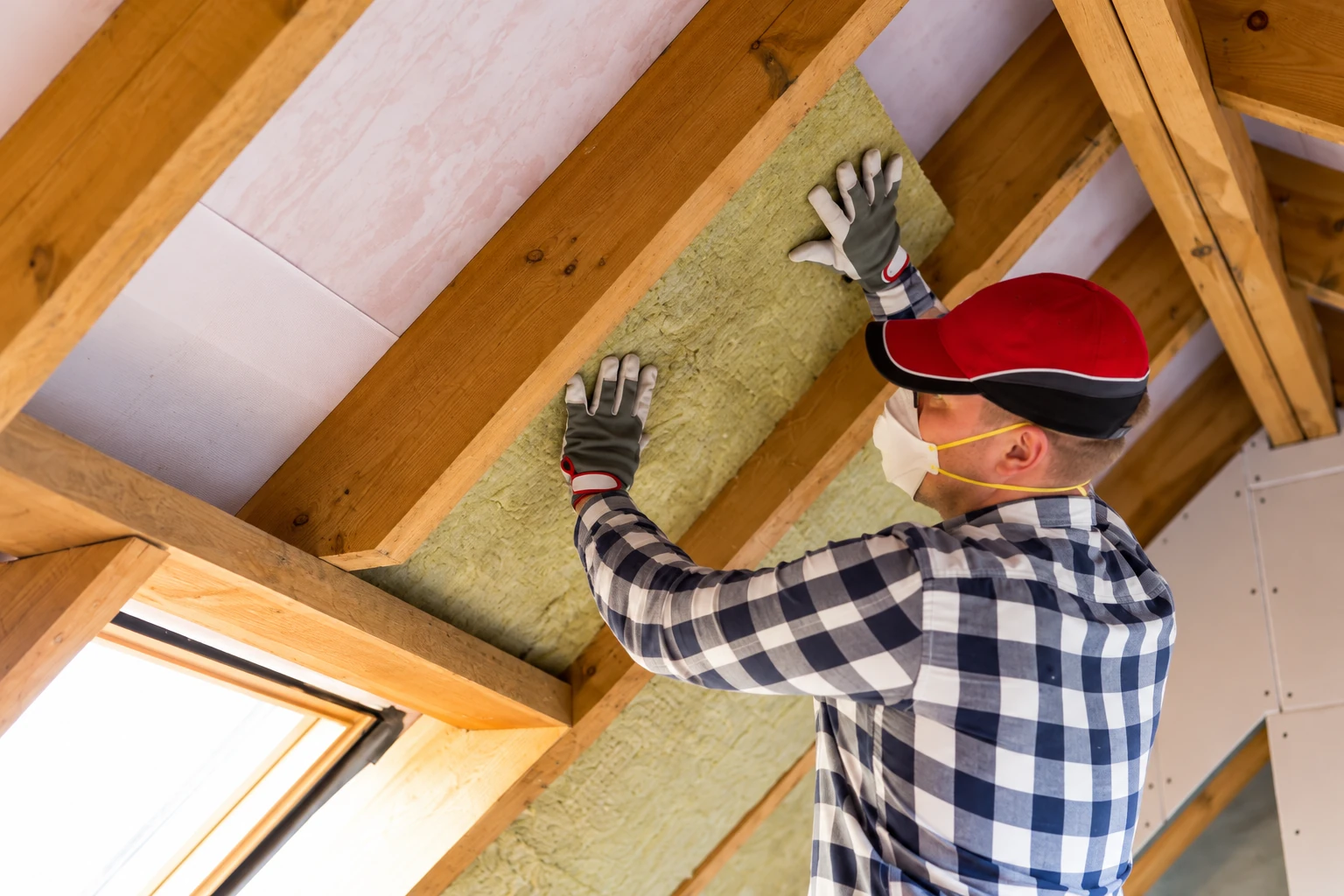 A worker in a red cap and respirator fits a yellow insulation batt between attic rafters.