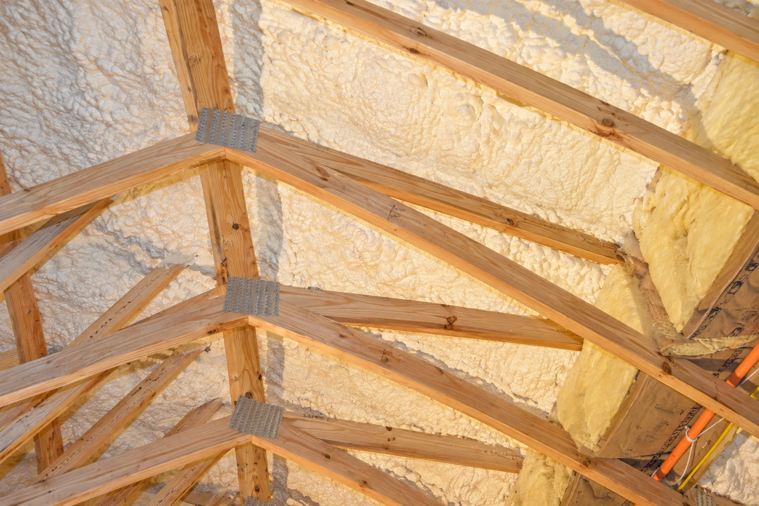 Cream spray foam coats the roof beneath exposed wooden trusses in an unfinished attic.