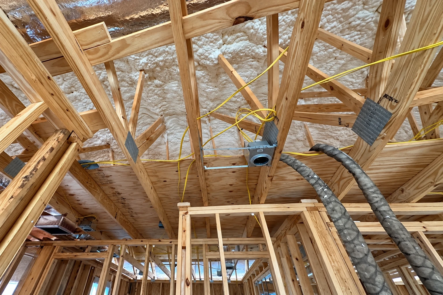 White foam insulation fills the roof framing of a commercial building, with yellow wiring and spray hoses visible.
