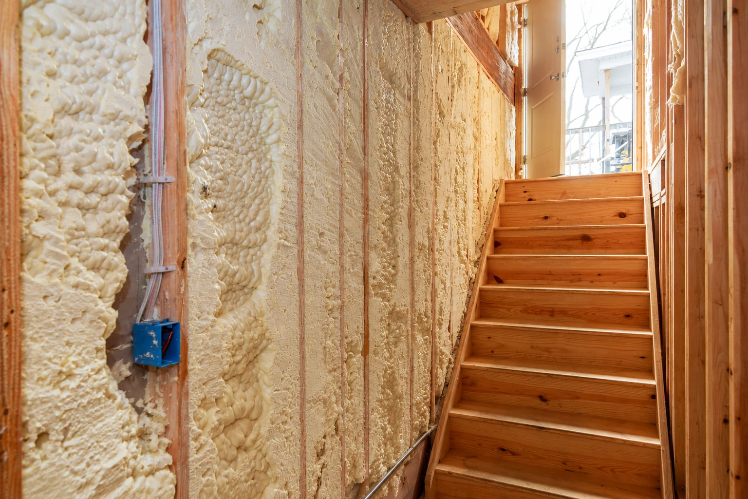 Exposed wall framing is densely filled with cream-colored spray foam beside a wooden staircase.