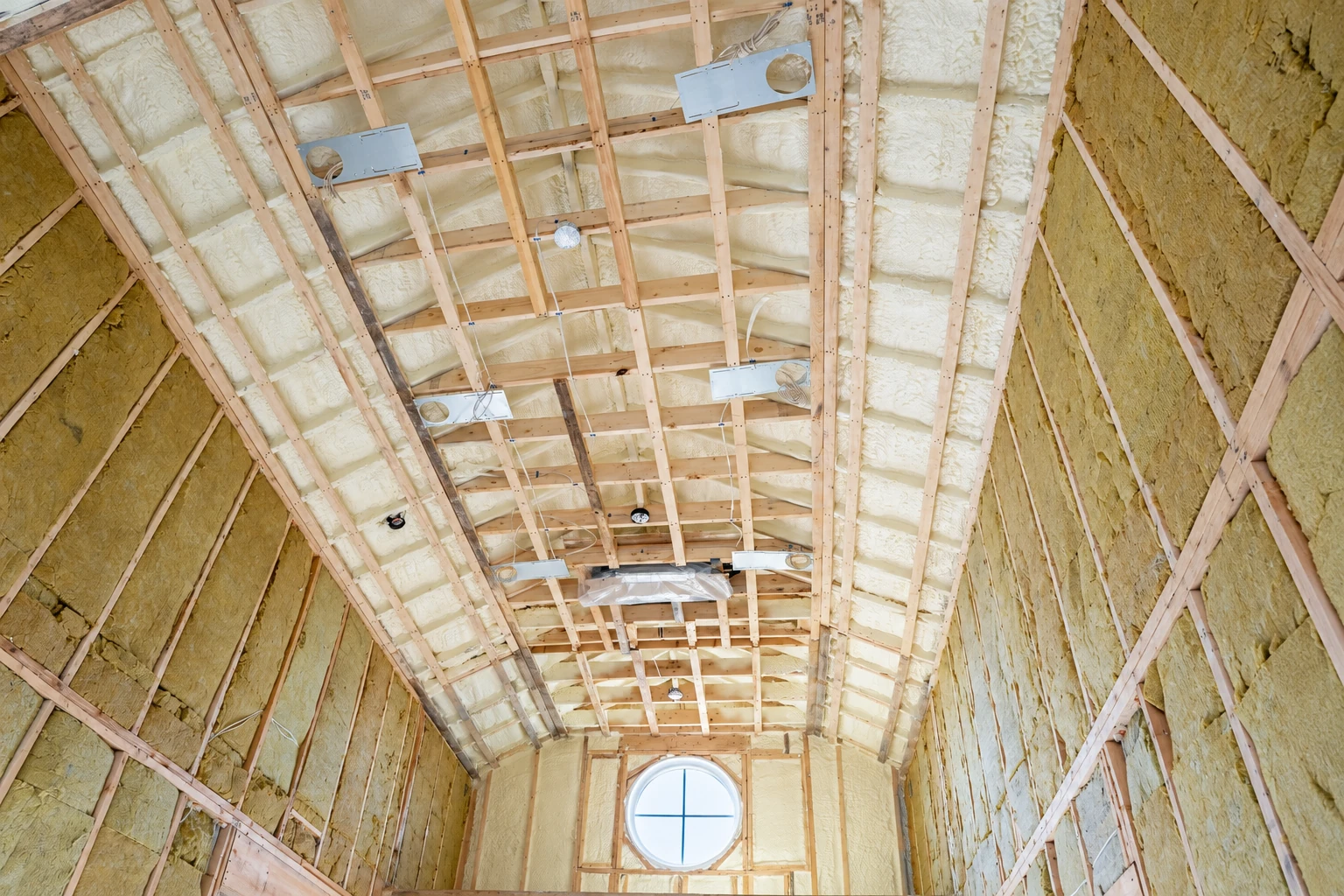 An unfinished vaulted interior shows exposed wooden framing with pale spray foam between the rafters and insulation on the side walls.